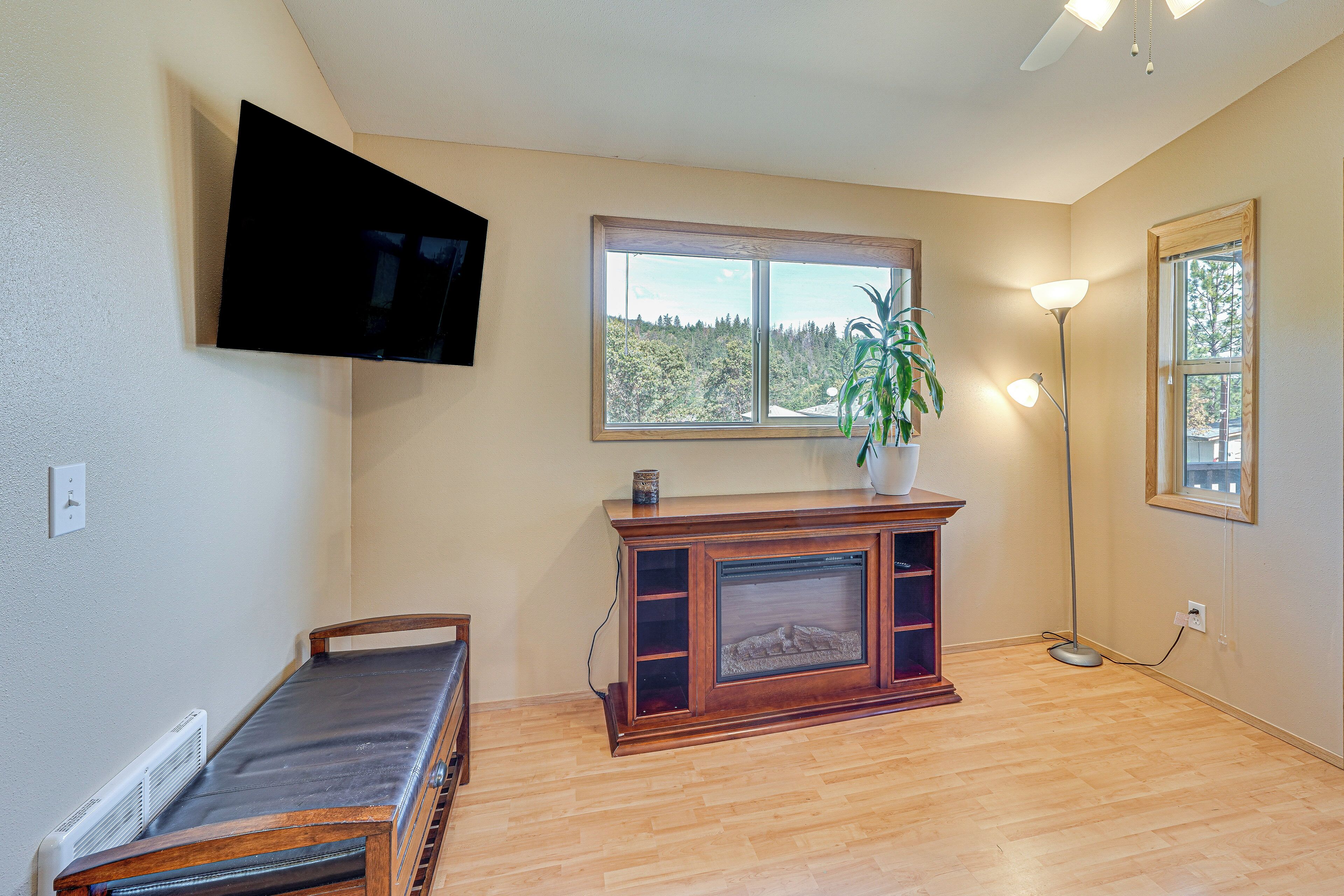 Una habitación con un banco de madera, un televisor montado en la pared, una ventana con vistas a árboles y una lámpara de pie.