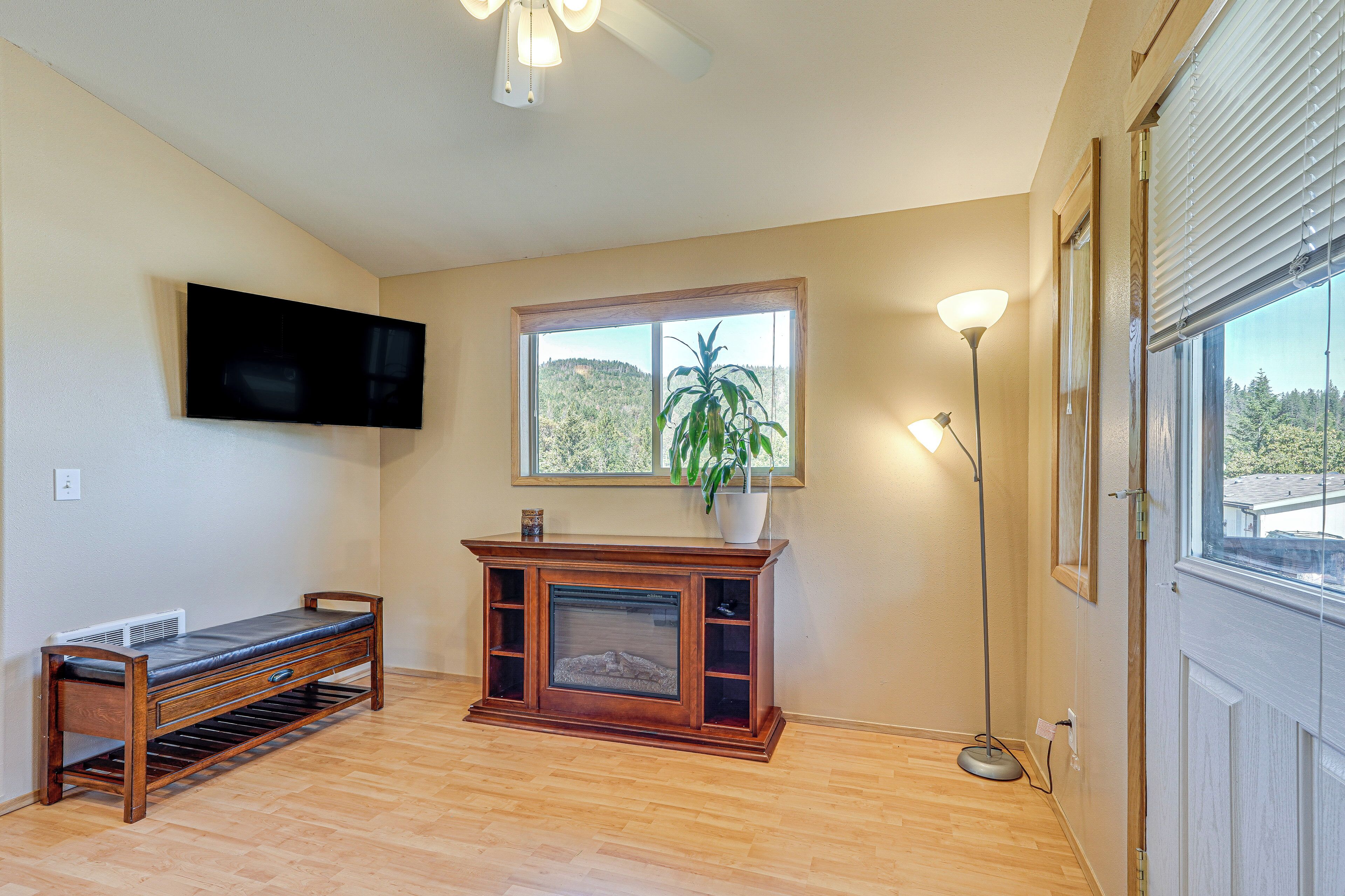 A living room with a wooden bench, a fireplace, a TV mounted on the wall, and a large window with a view of trees.