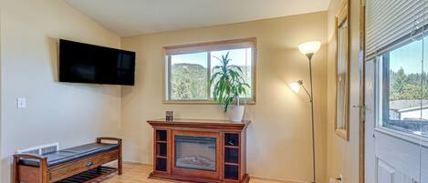 A living room with a wooden bench, a fireplace, a TV mounted on the wall, and a large window with a view of trees.