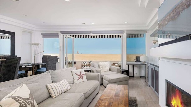 Een moderne woonkamer met een bank, een poef, een salontafel, een open haard en een groot raam met uitzicht op het strand.
