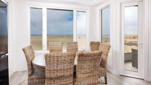 Een eethoek met rieten stoelen en een ronde tafel, grote glazen deuren met uitzicht op een strand.