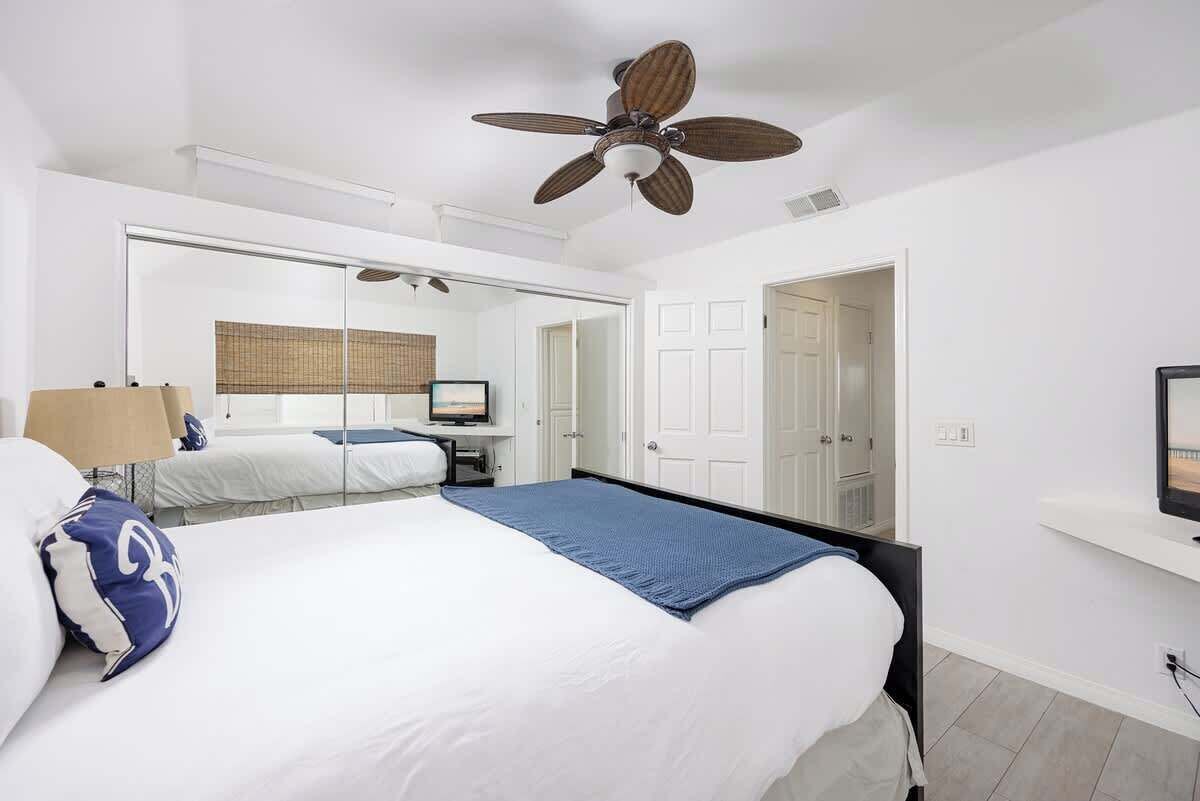 A bedroom with a bed, a ceiling fan, a mirror, a desk with a computer, and a door.