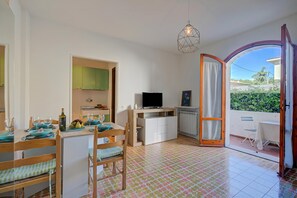 A room with a dining table set for two, a TV, a wooden chair, a small cabinet, and a view through an open door.