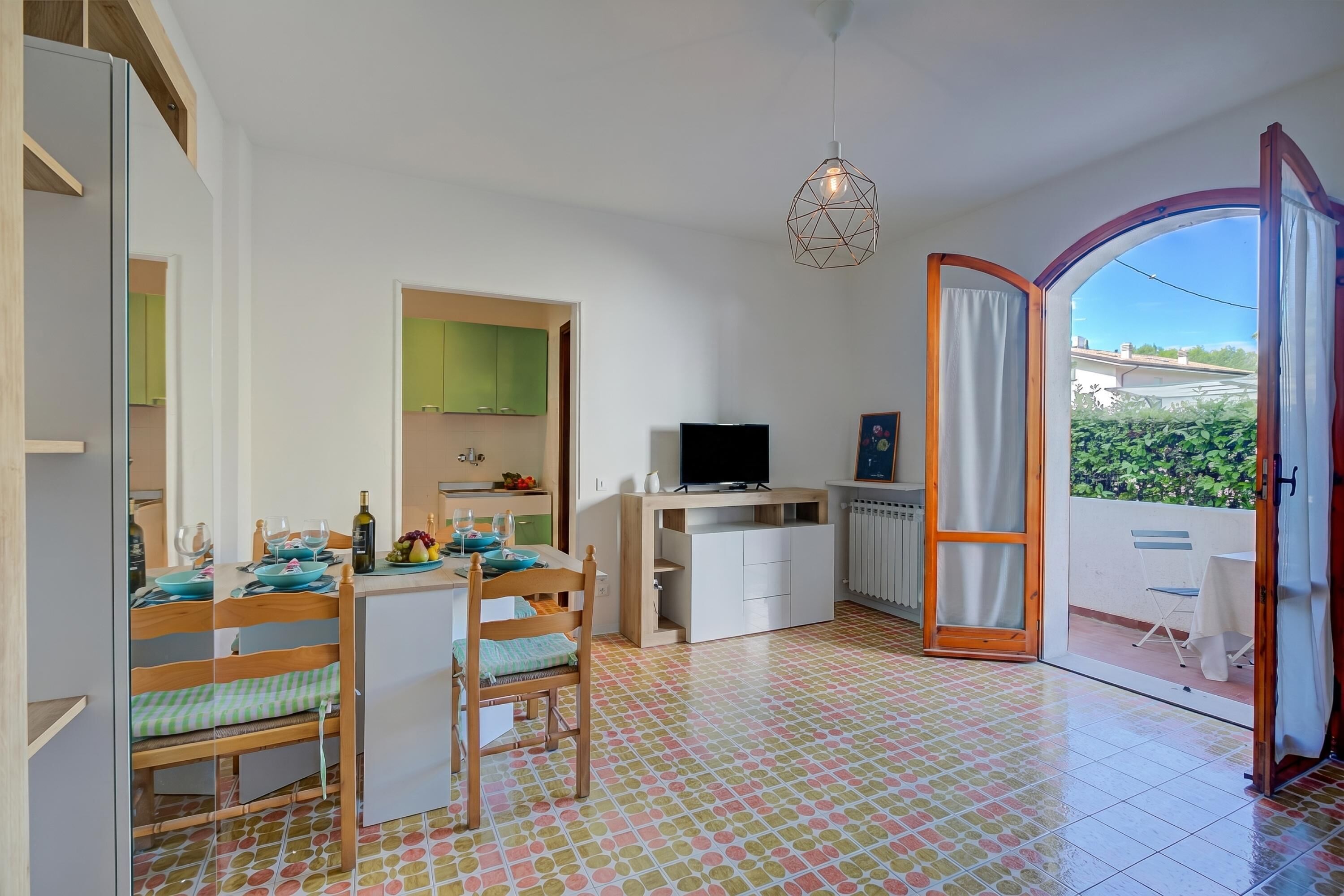 A room with a dining table, chairs, a TV, a small kitchen area, and a sliding glass door to an outdoor balcony.