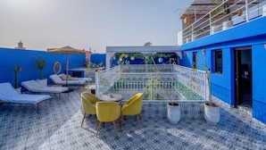Une terrasse sur le toit, avec des chaises longues, des chaises jaunes, des plantes en pot et un bâtiment bleu doté d’une balustrade blanche.