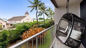Un balcón con una silla de mimbre, vista a una iglesia y vegetación tropical.