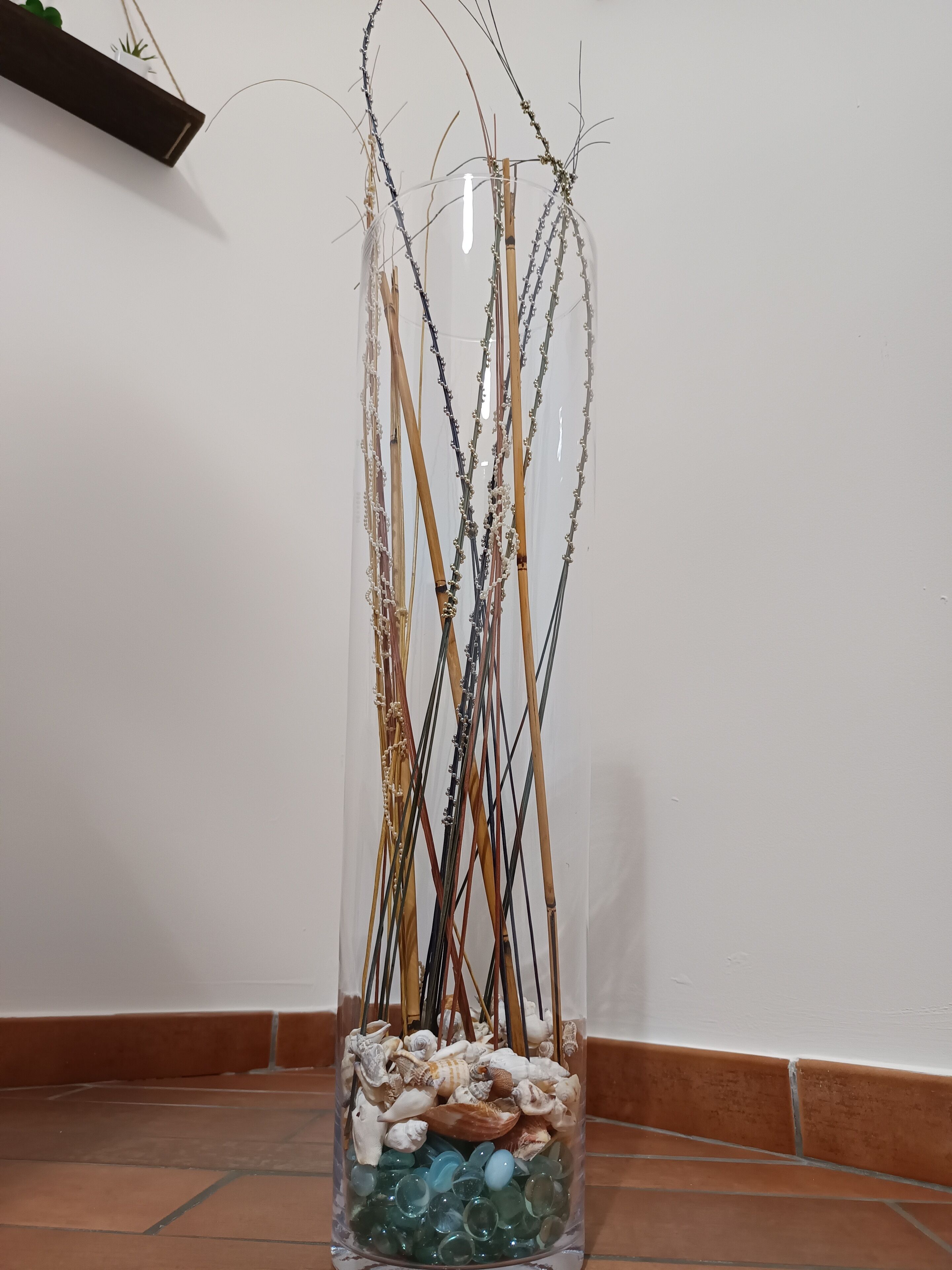 A tall, clear glass vase filled with various branches, seashells, and green stones, placed on a tiled floor against a plain wall.