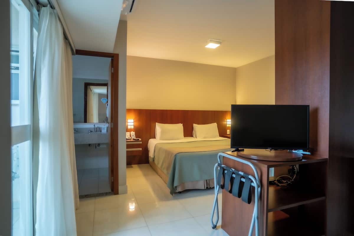 A hotel room with a bed, a TV, a bedside table with a lamp, and a bathroom visible through an open door.