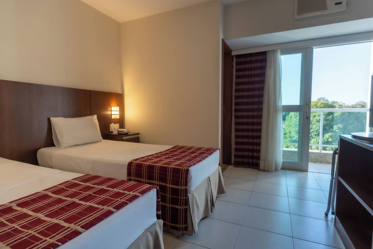 A hotel room with two beds, a nightstand, a lamp, a balcony with curtains, and a view of greenery outside.