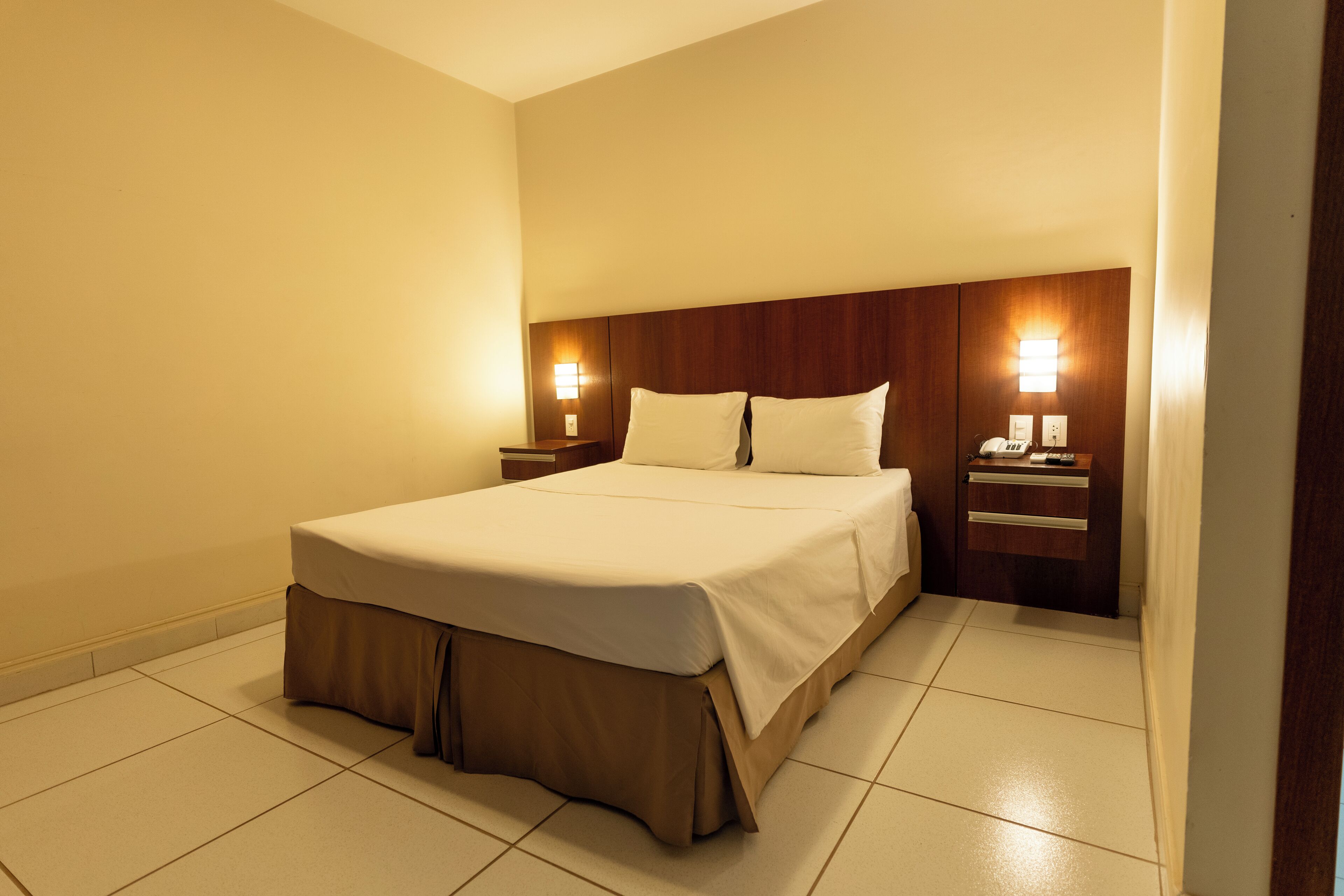 Une chambre d’hôtel avec un grand lit, une tête de lit en bois, des tables de chevet et des lampes fixées au mur.