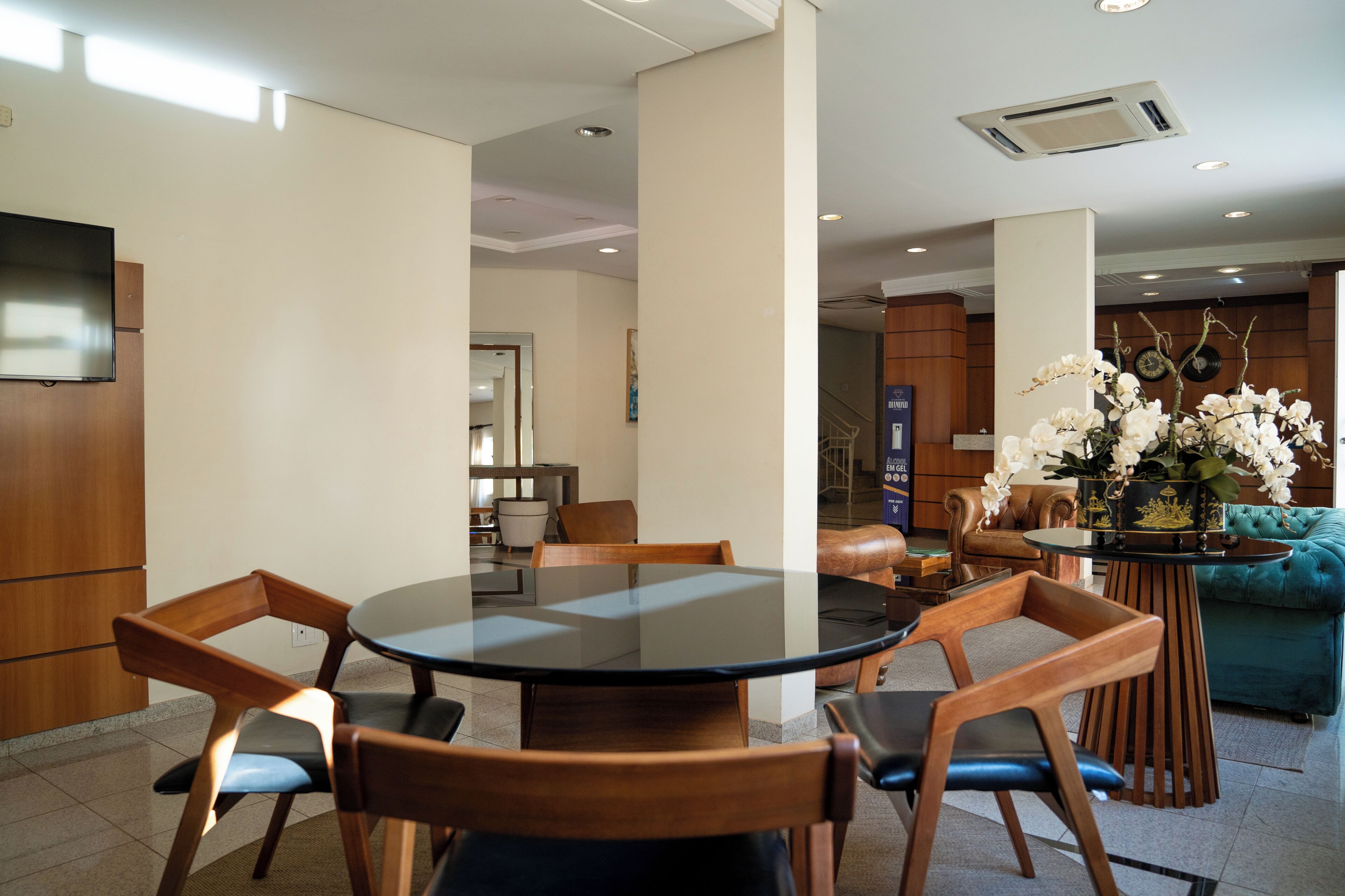 A modern hotel room with a round glass table, wooden chairs, a teal sofa, and a decorative orchid arrangement.