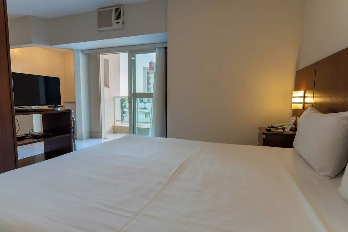 A hotel room with a large bed, a television on a desk, a bedside table with a lamp, and a view of the outside balcony.