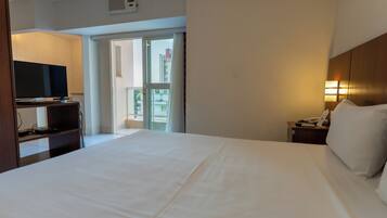 Habitación de hotel con una cama grande, un televisor sobre un escritorio, una mesita de noche con lámpara y vistas al balcón exterior.