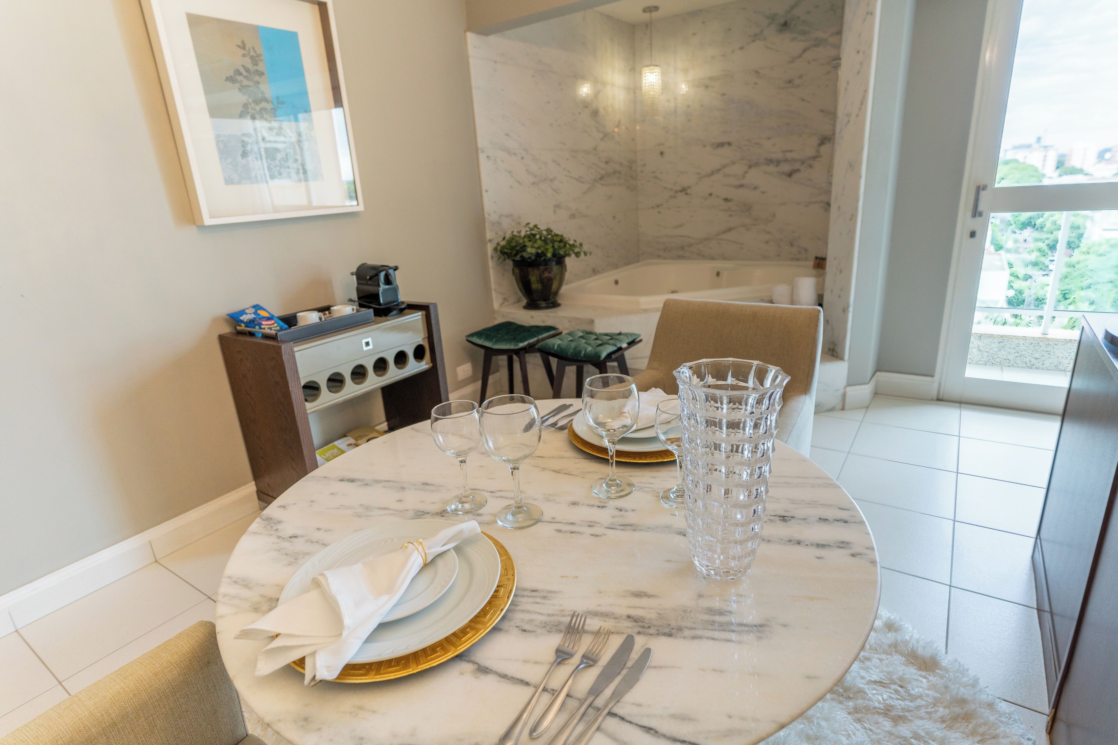A marble dining table set with plates, glasses, and cutlery, a coffee machine, and a potted plant in a modern room with a large window and a bathtub.
