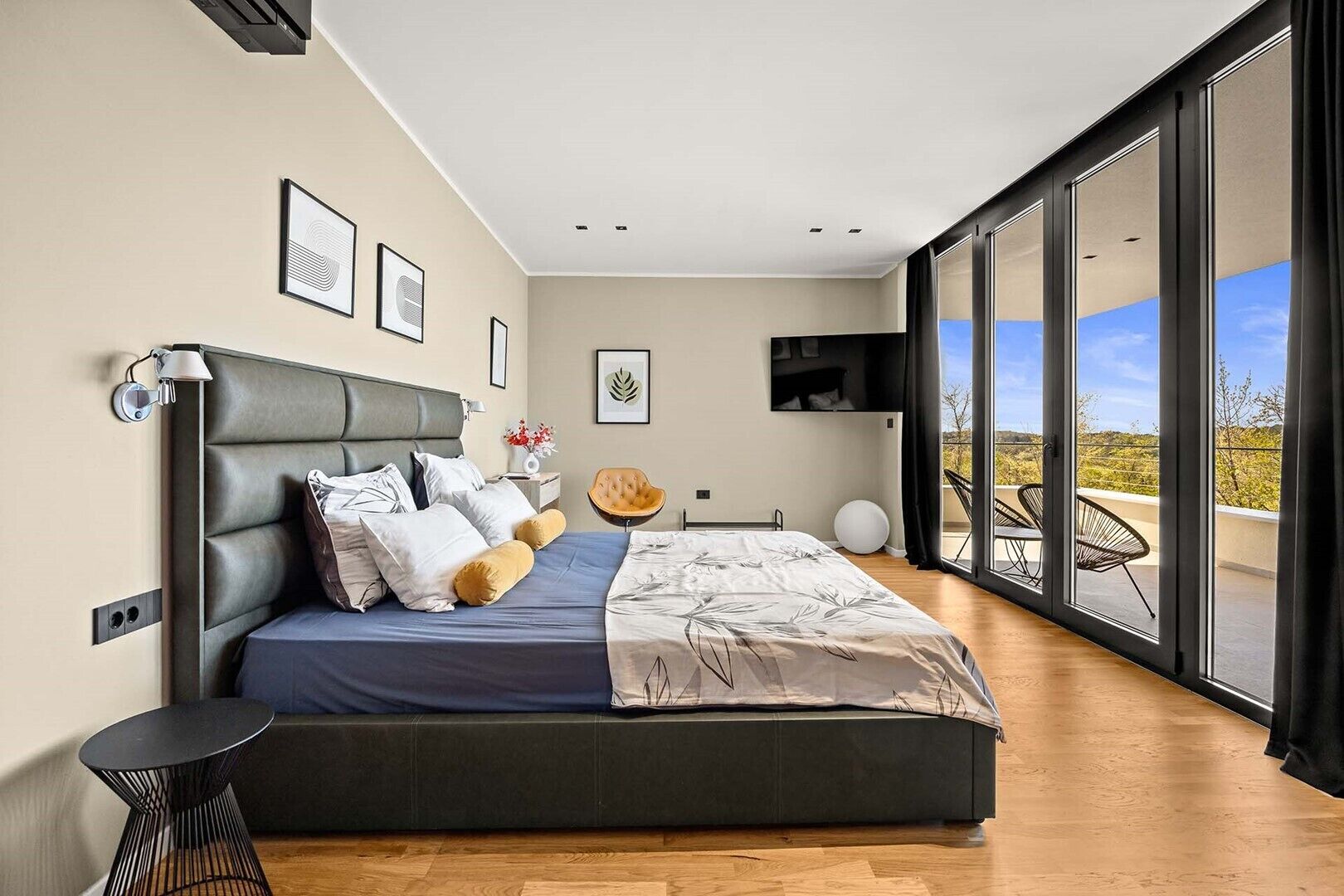 Een moderne slaapkamer met een groot bed, een televisie, een balkon met stoelen en uitzicht op de stad.