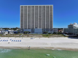 A high-rise hotel with multiple floors and balconies, situated next to a beach with sun loungers and a clear blue sky.