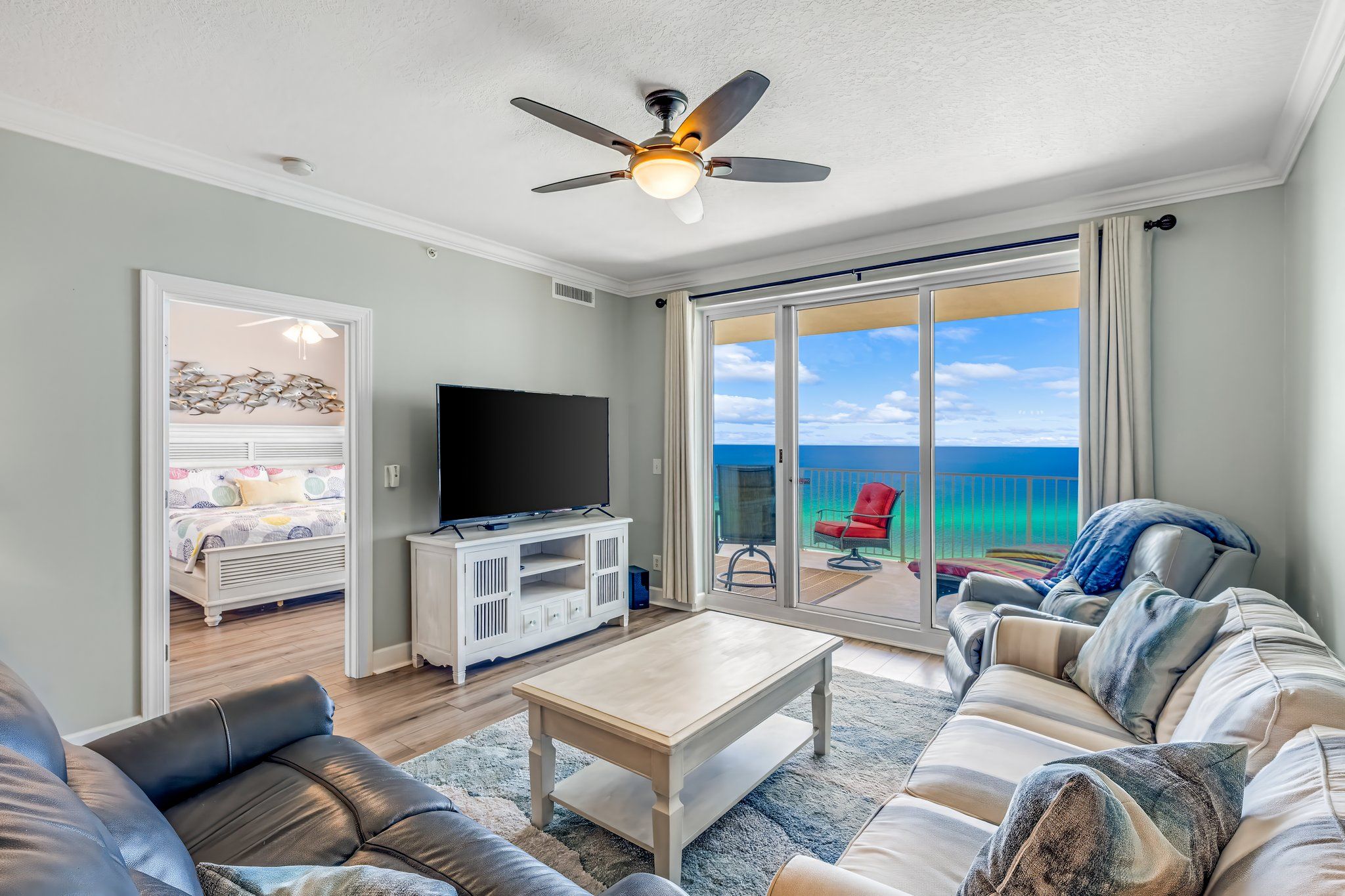 A spacious living room with a flat-screen TV, a ceiling fan, and a large sliding glass door leading to a balcony with a view of the ocean.