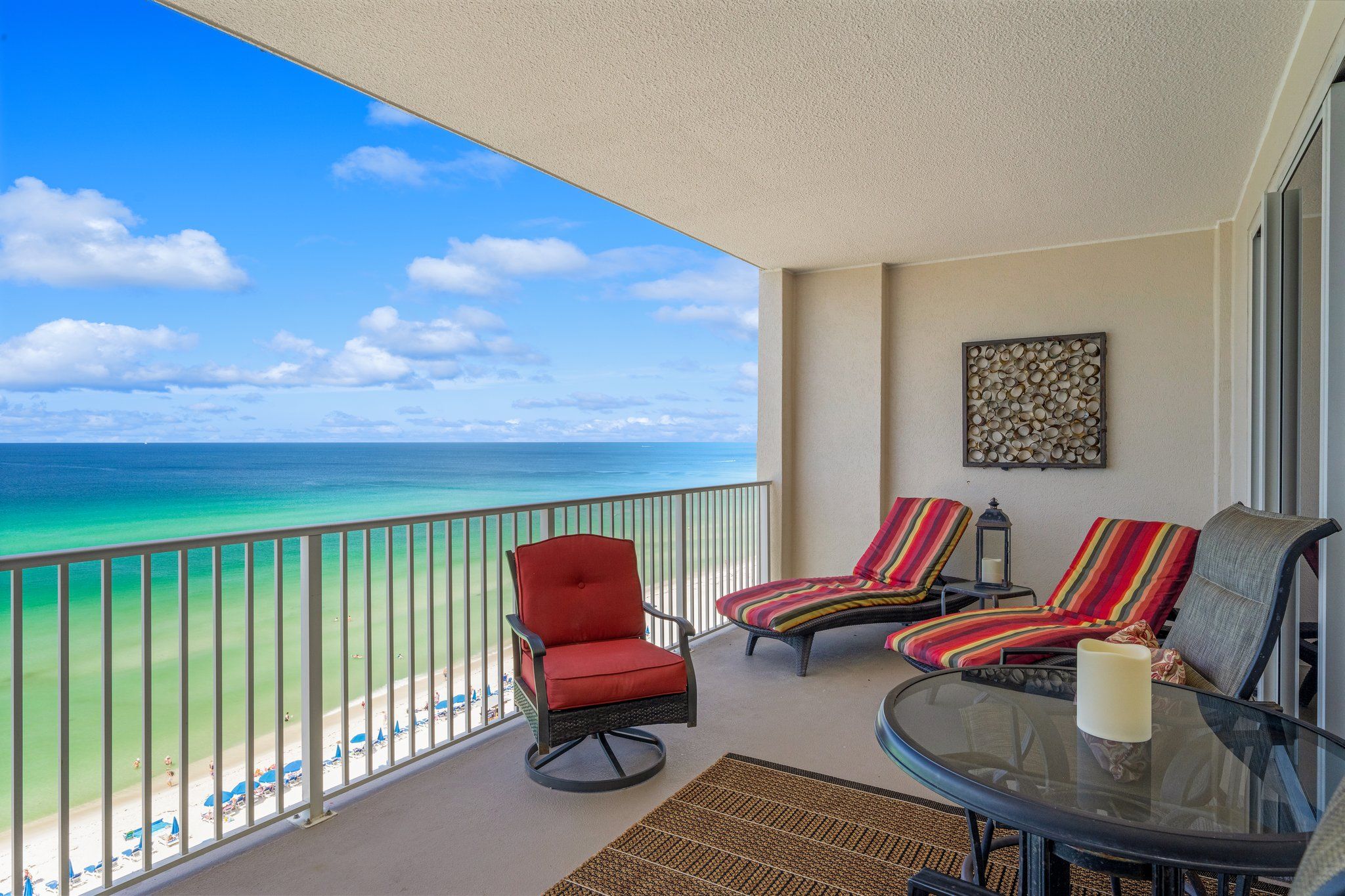 A balcony with a view of the ocean, equipped with a glass table, two striped lounge chairs, and a red chair.