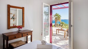 A bedroom with a bed, a wooden dresser, a mirror, and a view of the sea through an open door.