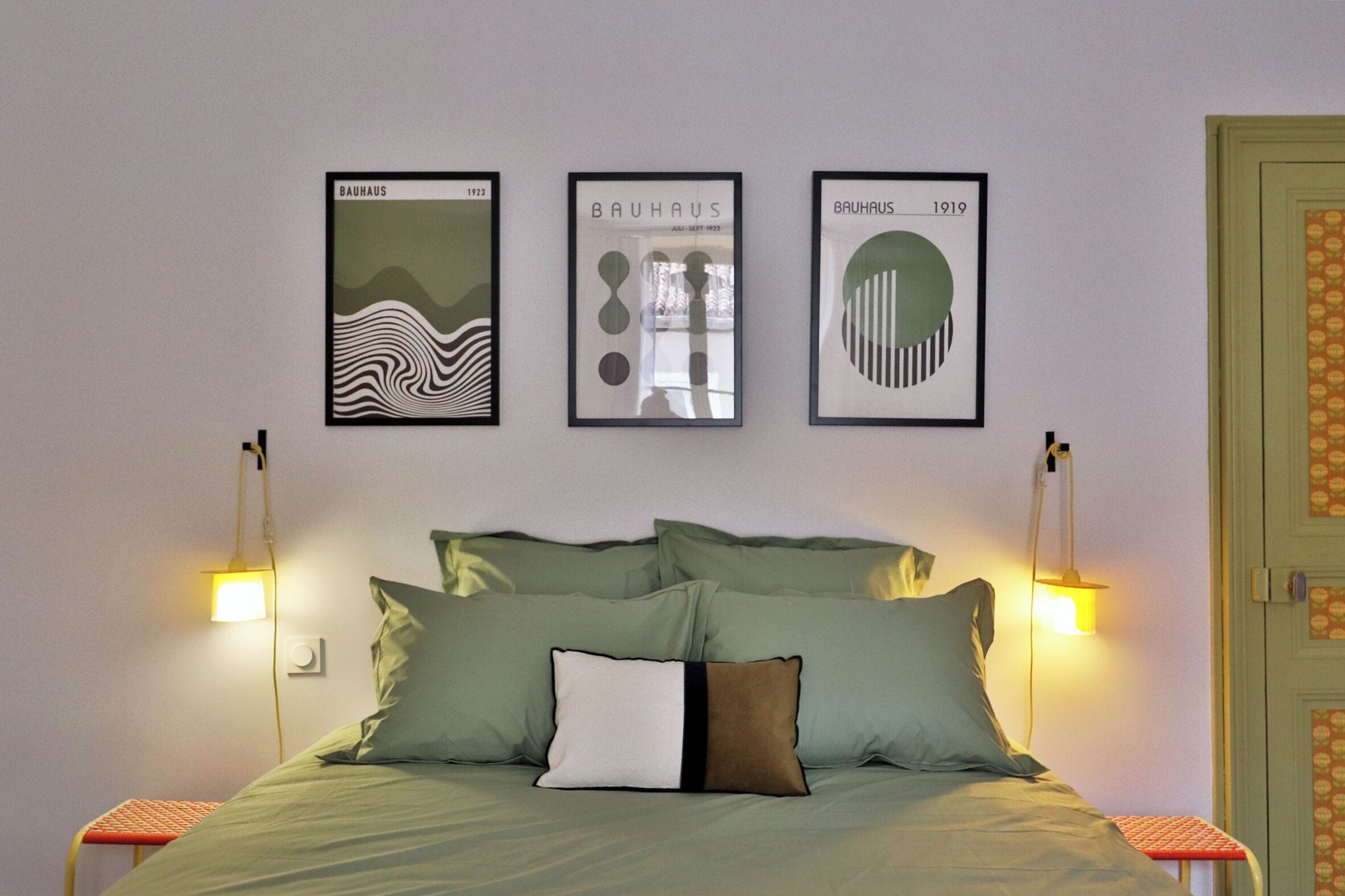 Ein Bett mit grüner Bettwäsche, ein Kopfkissen mit schwarzem Streifen und drei gerahmte Kunstwerke an der Wand.