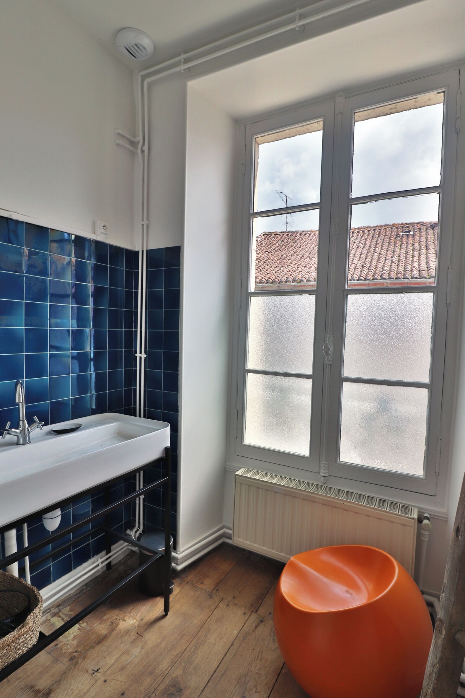 Un baño con un lavabo blanco, azulejos azules, un ventanal con vidrio esmerilado y suelo de madera.