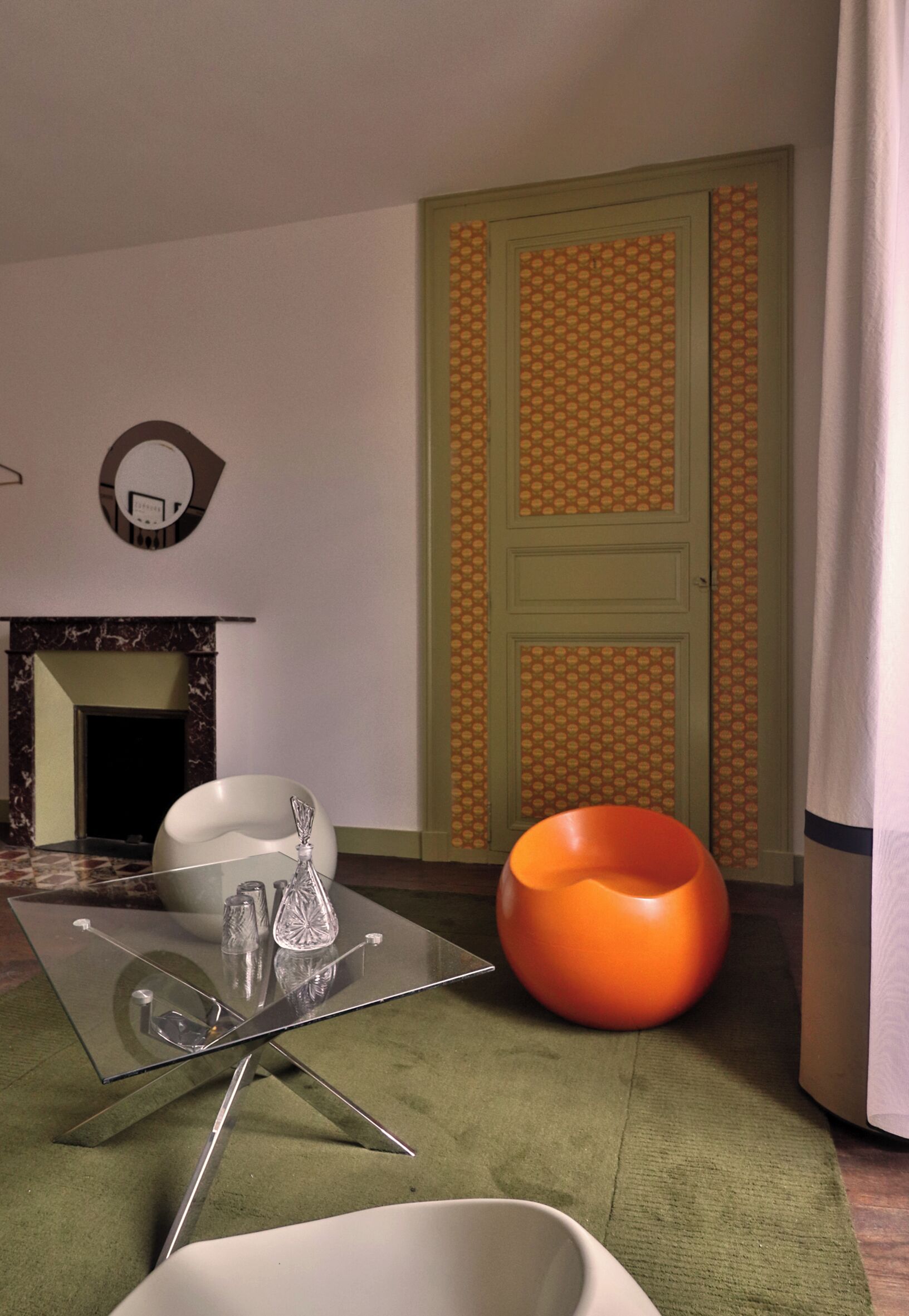 Una habitación moderna con una mesa de vidrio, una silla blanca, un sillón de peluche naranja y una puerta verde.