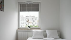 Un dormitorio con cama, ropa de cama blanca, una ventana con persianas y un radiador.