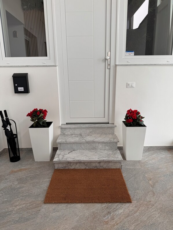 Une porte blanche avec un trou de serrure, une boîte aux lettres et deux plantes en pot, chacune ornée de fleurs rouges, de part et d’autre.
