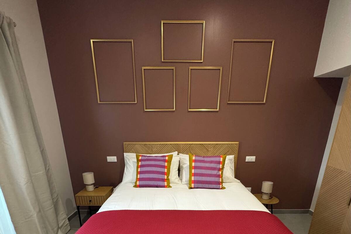 Una cama con una colcha roja, almohadas blancas y un cabecero de madera. Se observan interruptores de luz en la pared y una pequeña mesa con una lámpara junto a la cama.