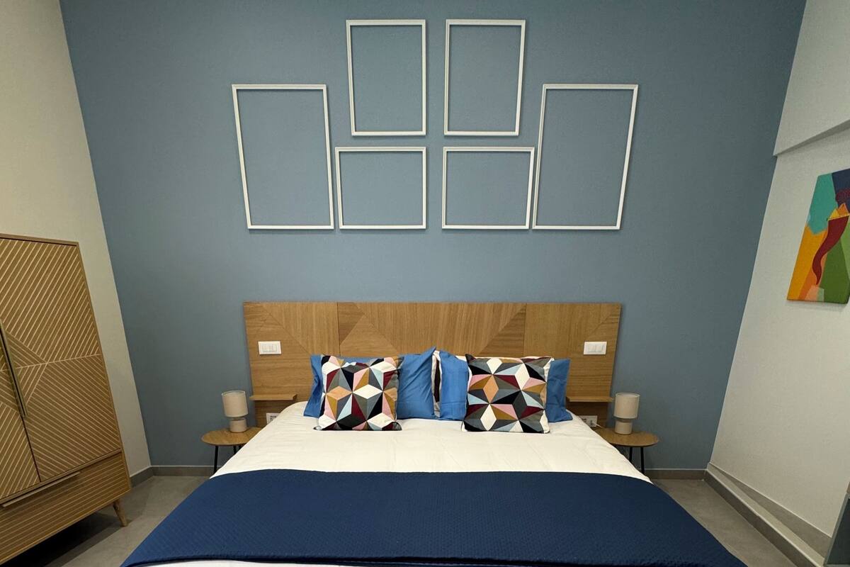 Un dormitorio moderno con cabecera de madera, una cama grande con una manta azul, varios cojines y una pared decorada con motivos geométricos.