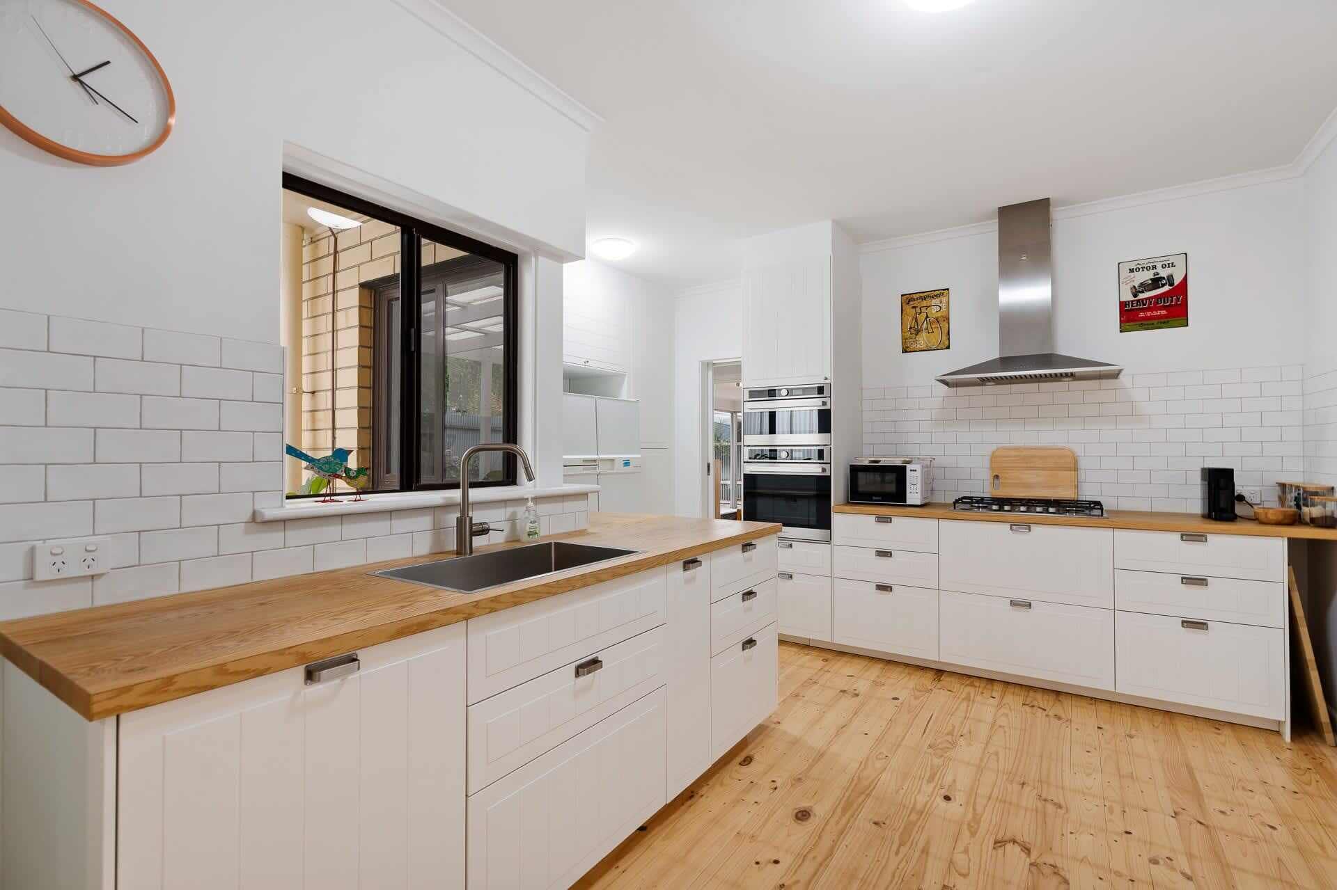 A modern kitchen with white cabinetry, a wooden countertop, a stainless steel range hood, and a window with blinds.