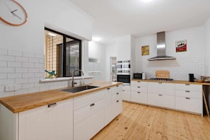 Une cuisine moderne avec des meubles blancs, un plan de travail en bois, une hotte en acier inoxydable et une fenêtre équipée de stores.