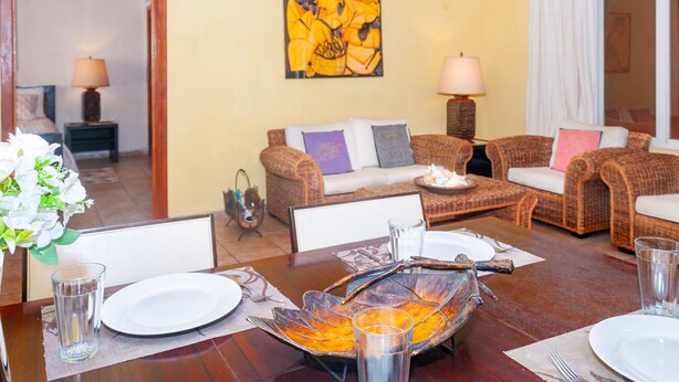 Un coin repas avec une table en bois dressée pour deux personnes, un centre de table décoratif en verre et un canapé avec des coussins en arrière-plan.