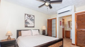 Une chambre à coucher comprenant un lit, des tables de chevet, un ventilateur de plafond, un appareil de climatisation et une salle de bain visible à travers une porte ouverte.