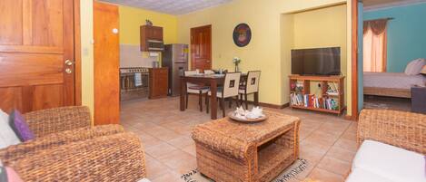 A living room with wicker furniture, a dining area with a table and chairs, a kitchen with appliances, and a bedroom with a bed and curtains.