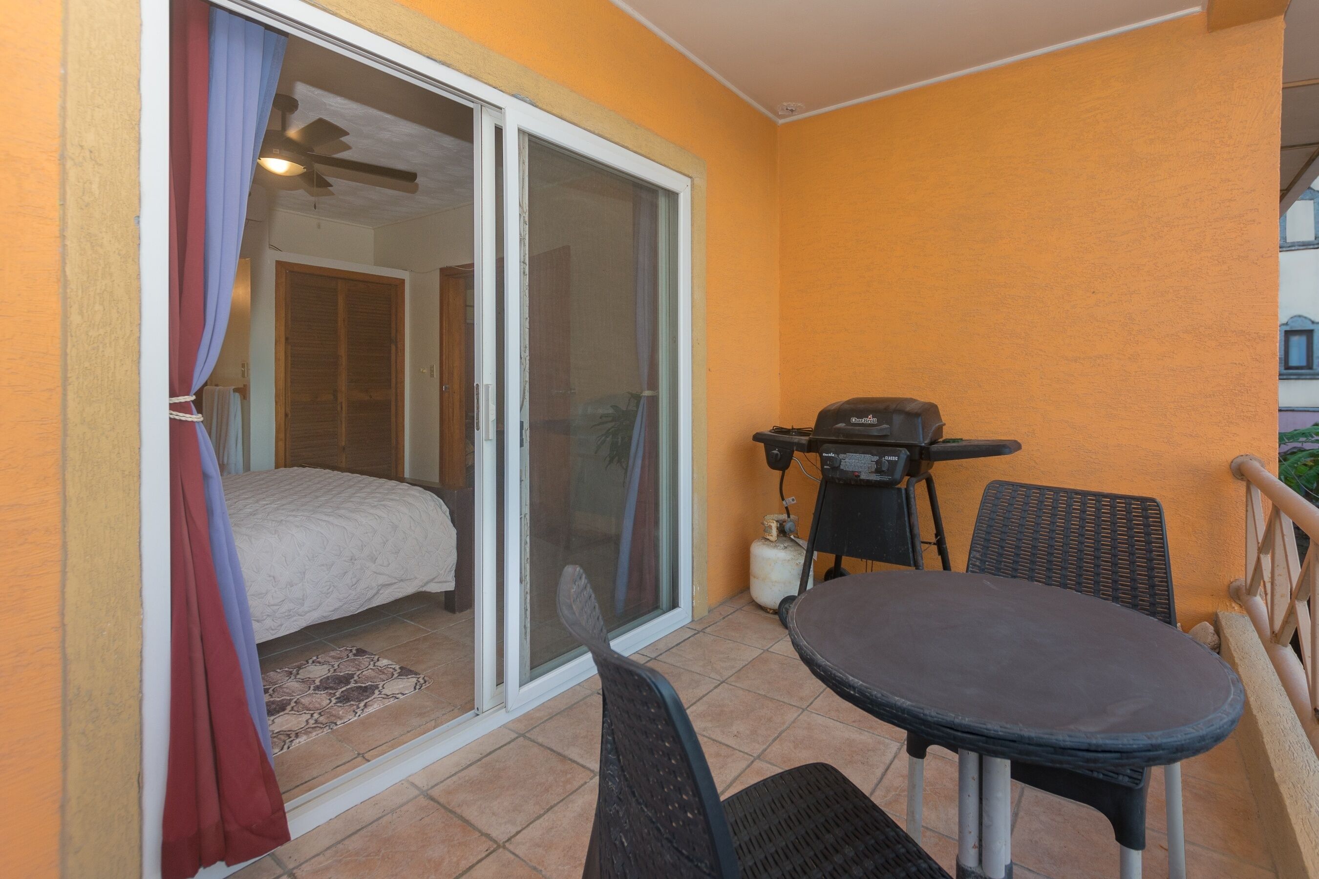 A balcony with a sliding glass door, a small table with chairs, a barbecue grill, and a gas cylinder.