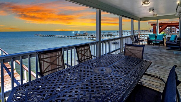 A covered outdoor seating area with a view of the ocean and a pier in the distance during sunset. - Shore Better Than Work 4 Bedroom Home by RedAwning (Rockport)