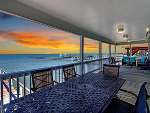 A covered outdoor seating area with a view of the ocean and a pier in the distance during sunset. - Shore Better Than Work 4 Bedroom Home by RedAwning (Rockport)