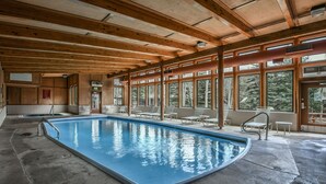 An indoor swimming pool with wooden beams, large windows, and lounge chairs.