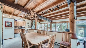 A rustic wooden interior with a dining table and chairs, a stone pillar, and a view of the outdoors through large windows.