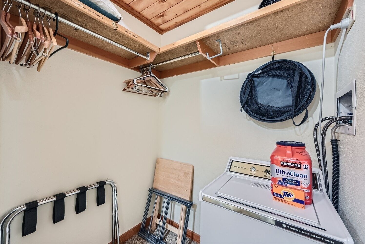 A compact laundry room with a front-loading washer and dryer, a hanging clothes rack, a portable vacuum cleaner, and a shelf with a bottle of detergent.