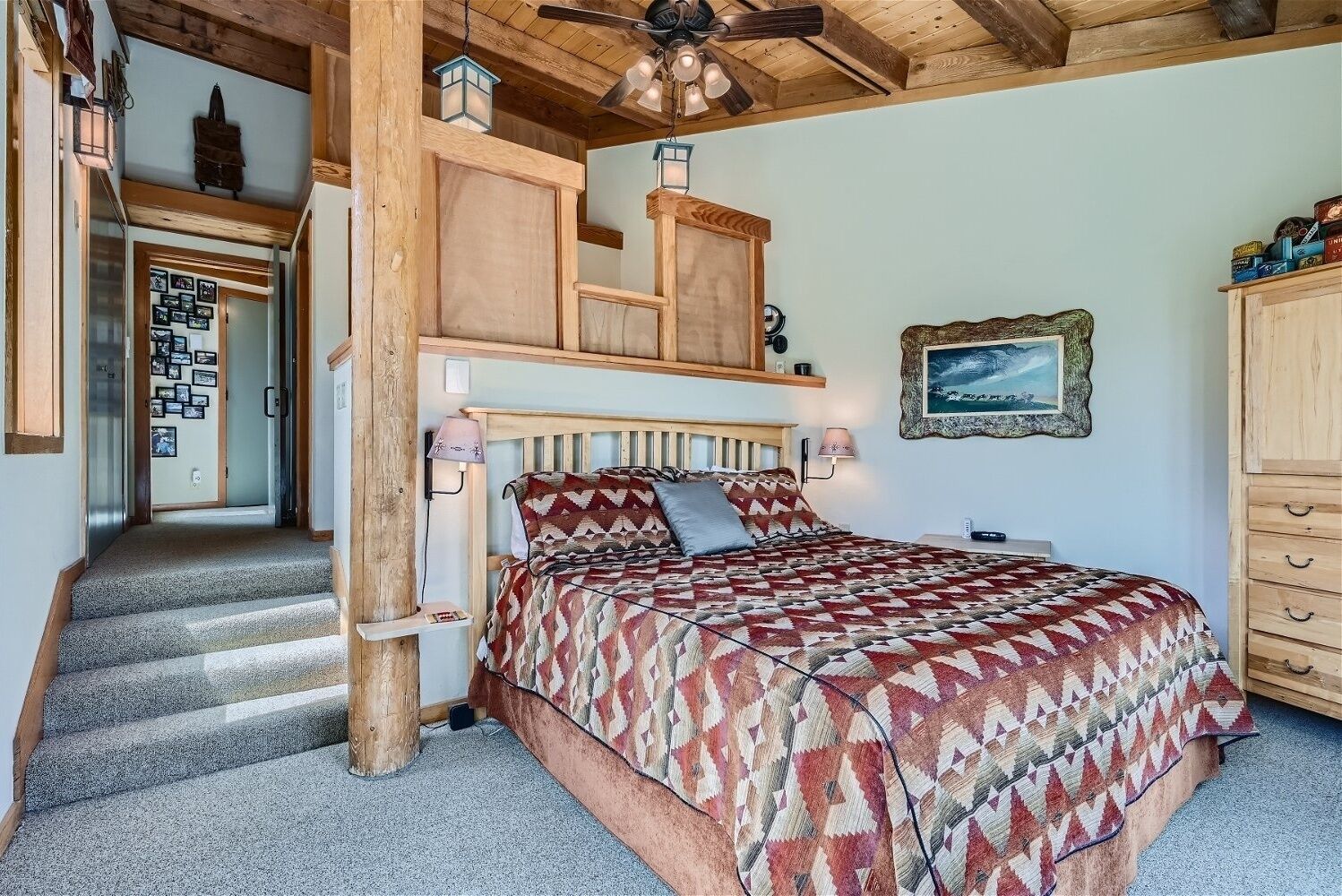Une chambre à coucher avec un lit, une tête de lit en bois, une table de chevet, une lampe, un tableau au mur et un escalier menant à une autre pièce.