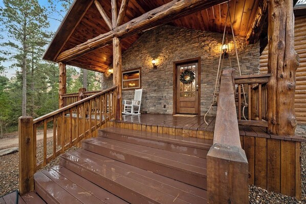 A wooden cabin with a stone exterior, a porch with wooden railings, and a white rocking chair. - The Rustic Ritz|2BD 2BTH|Hot tub|Shuffleboard|Luxury|SLPs8 (Broken Bow)