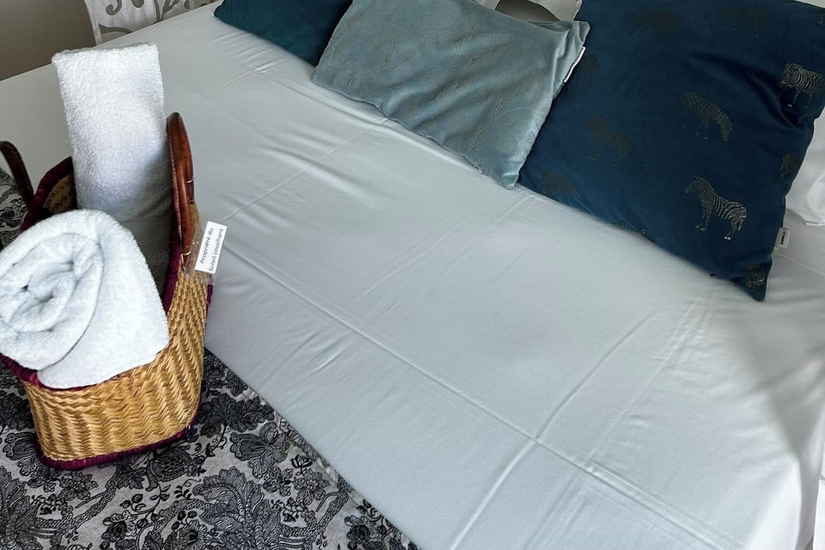Una cama bien hecha con ropa de cama blanca y verde azulado, un cuadro decorativo en la pared y una cesta de mimbre con una toalla.