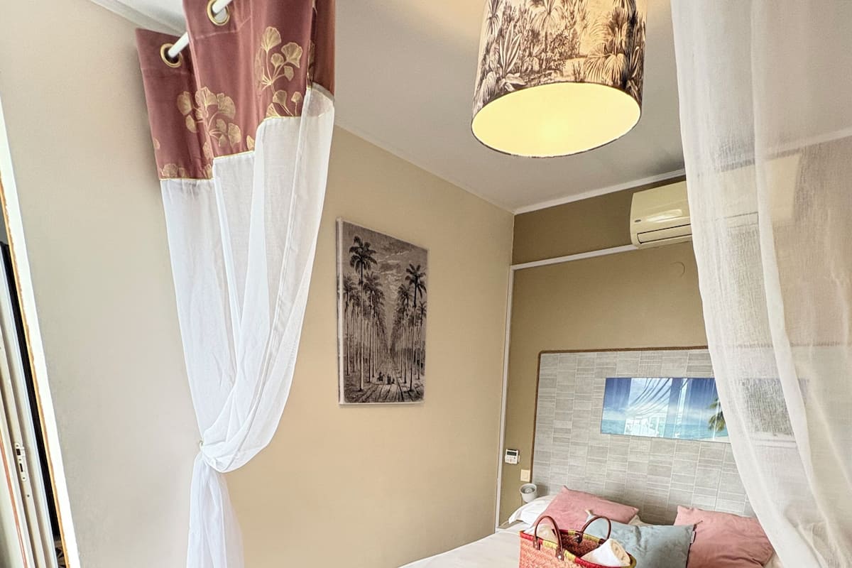 Un dormitorio con cama, banco, cortinas, una obra de arte en la pared y una lámpara de techo.
