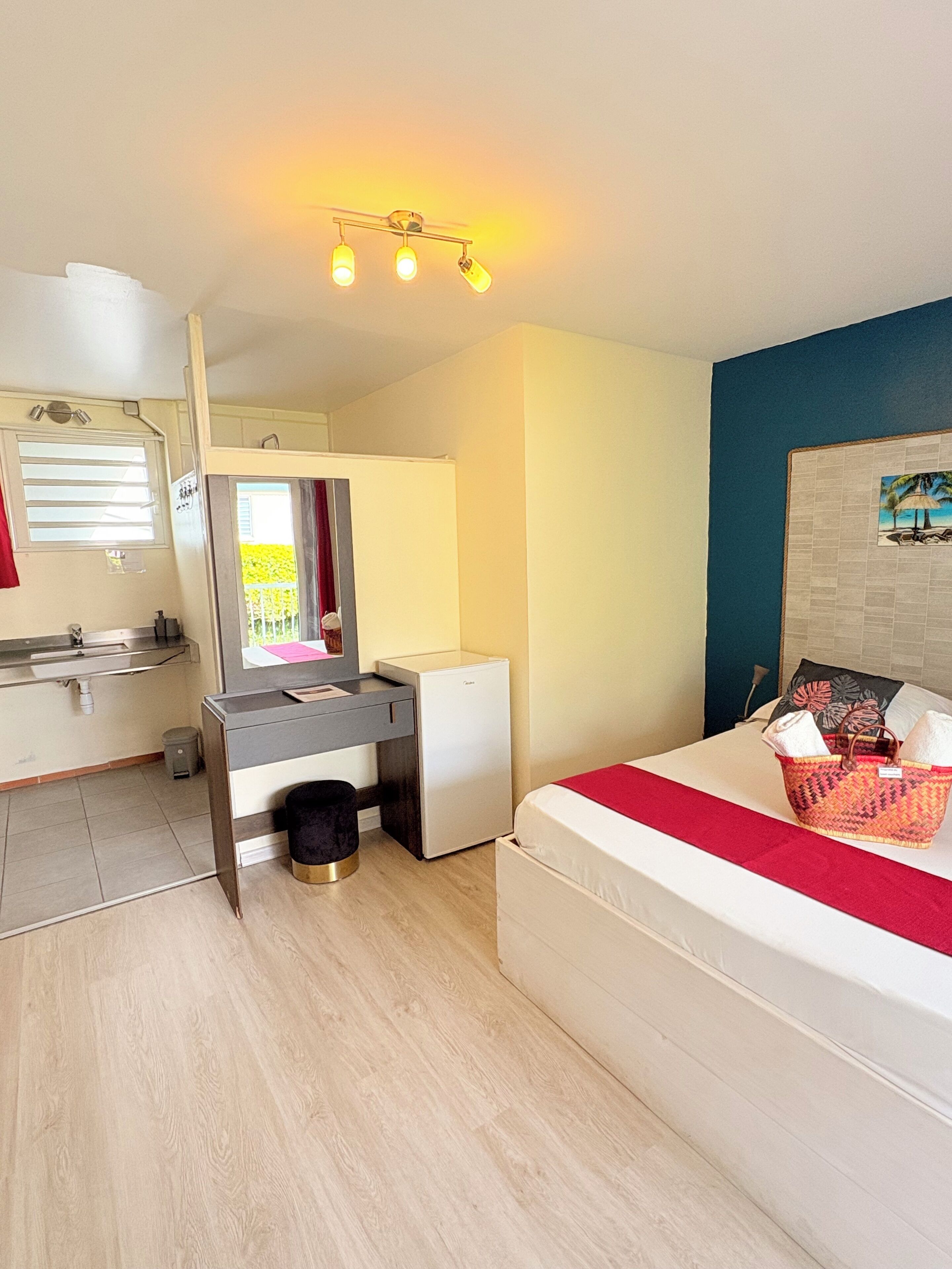 A compact room with a bed, a desk, a small refrigerator, a sink, and a window with curtains.