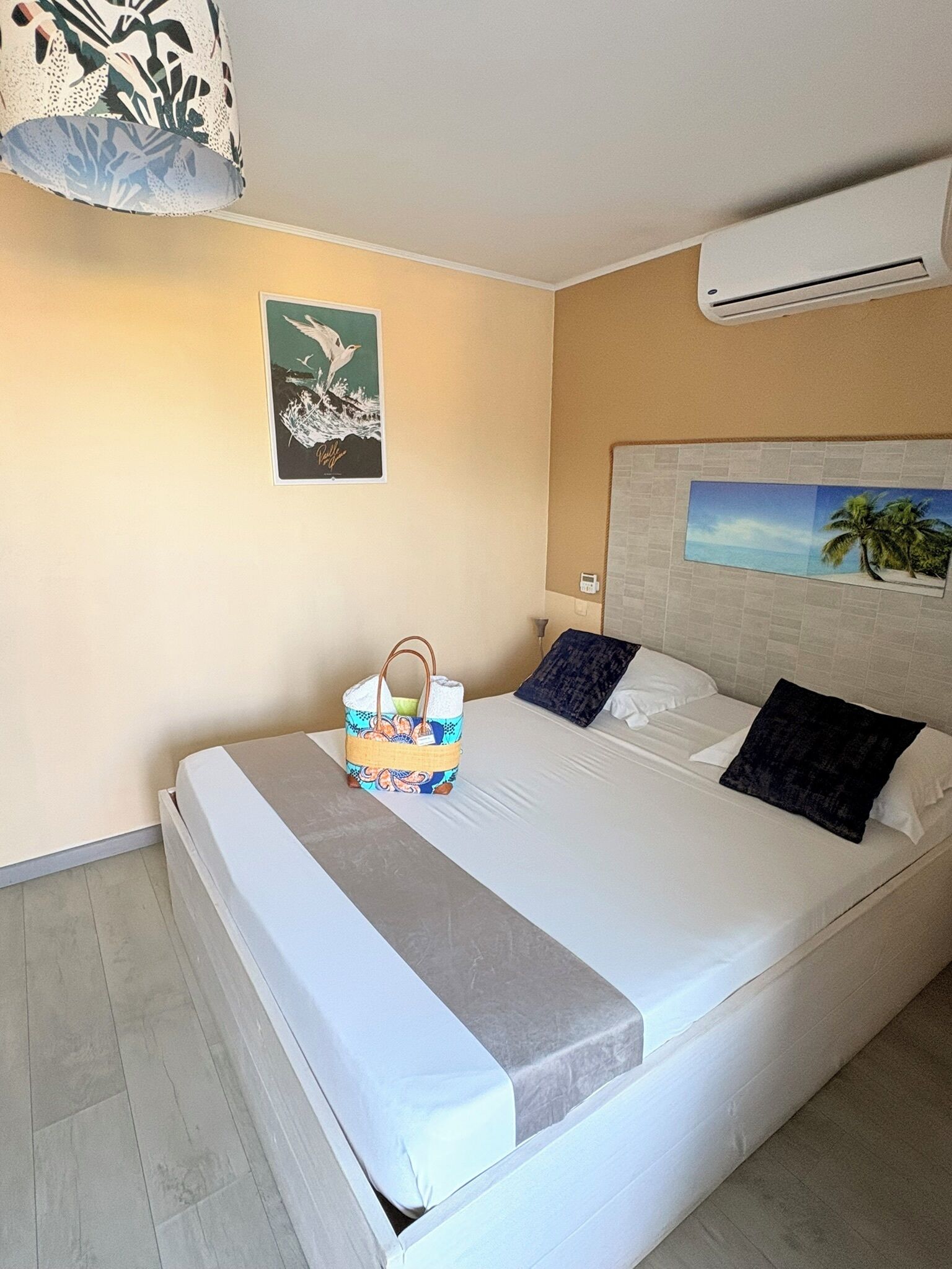 Una habitación de hotel con una cama, aire acondicionado de pared, un cuadro enmarcado y una mesita con una cesta.