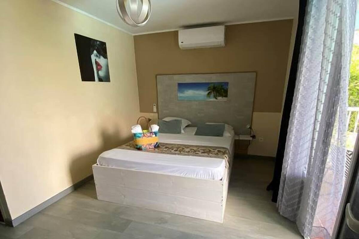 Un dormitorio con cama, ventilador de techo, unidad de aire acondicionado, un cuadro en la pared y una ventana con cortinas.