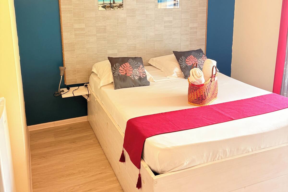 Un dormitorio con cama, almohadas, cabecero con obra de arte, mesita de noche y una manta roja.