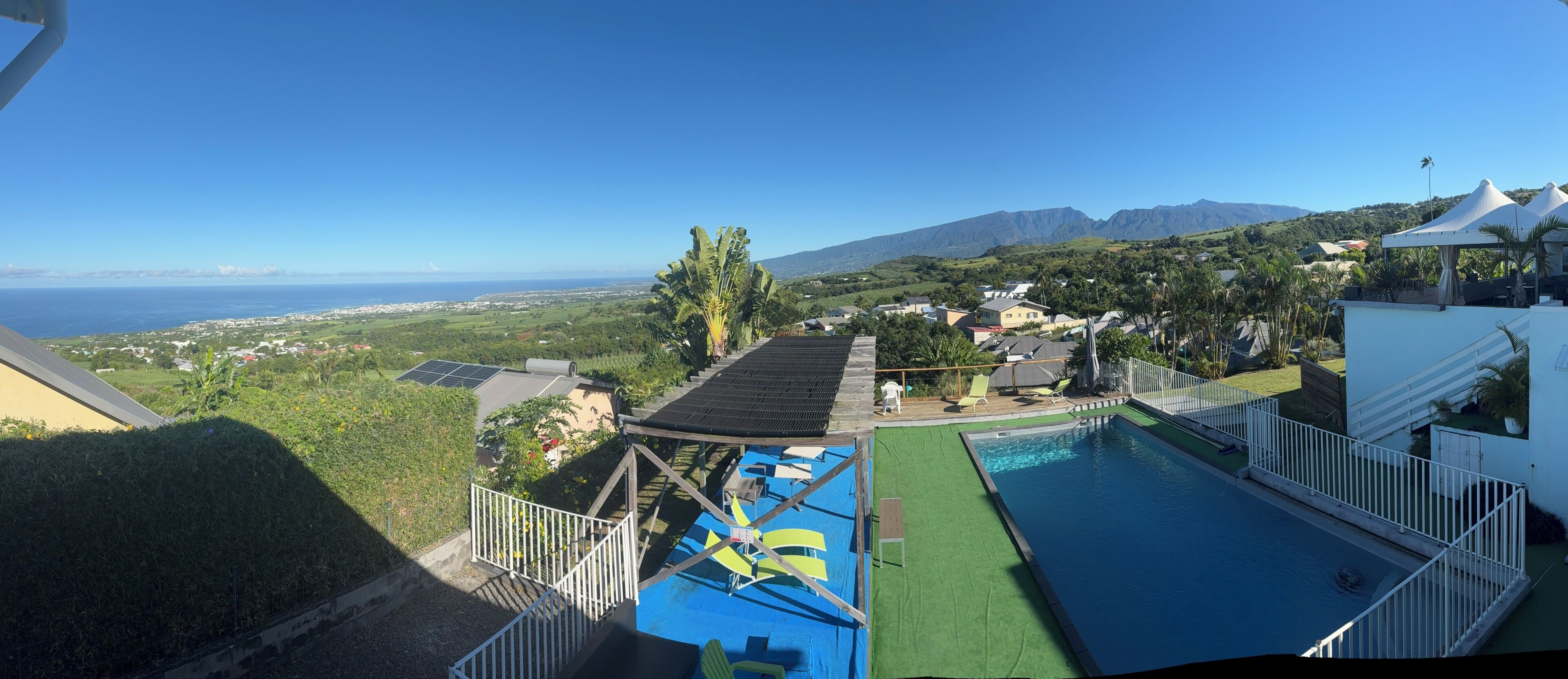 Un espace piscine offrant une vue sur l’océan et les montagnes, une clôture blanche et un bâtiment doté d’un toit en pente.